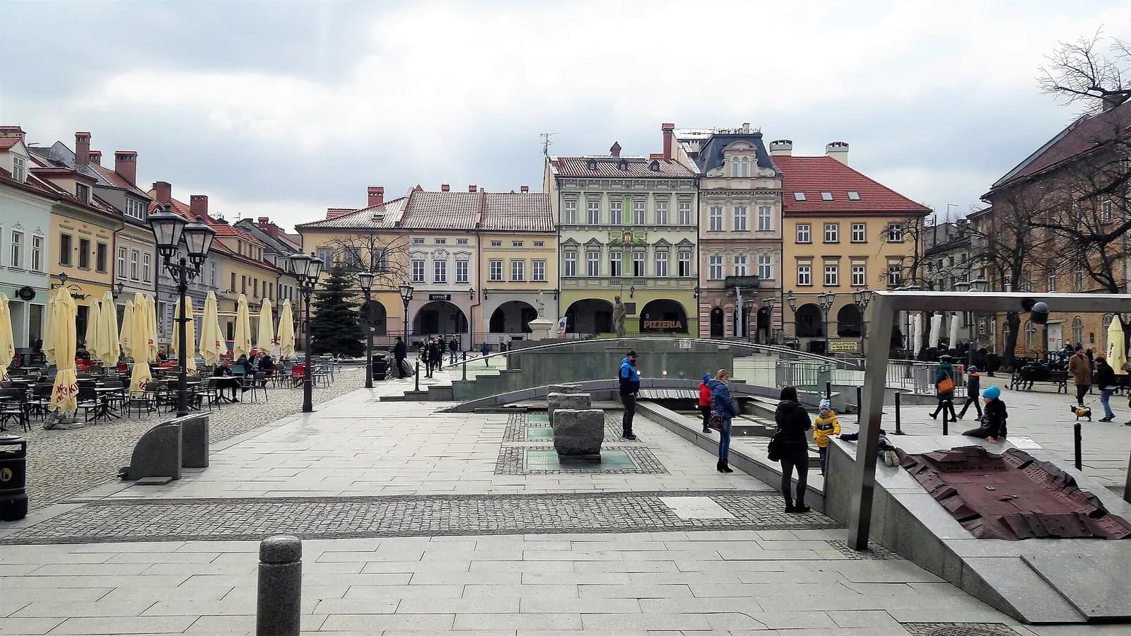 A wide view of a European town square with historical buildings lining the background, people walking around, and an outdoor cafe with yellow umbrellas on the left.